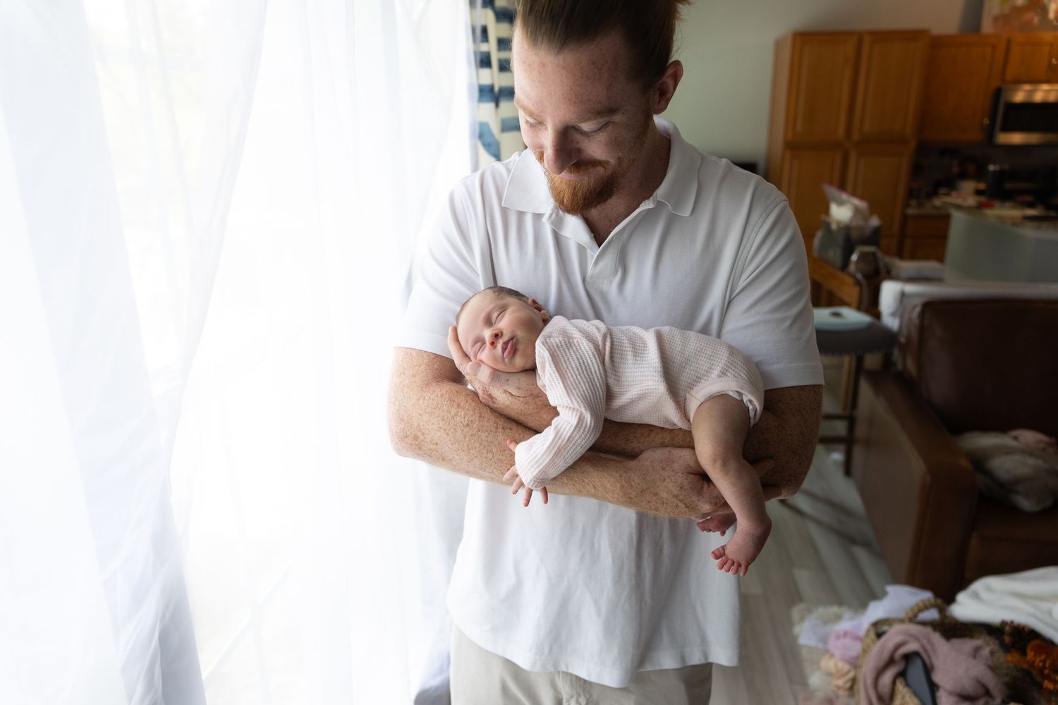 Parent holding sleeping newborn baby near bright window in home setting.
