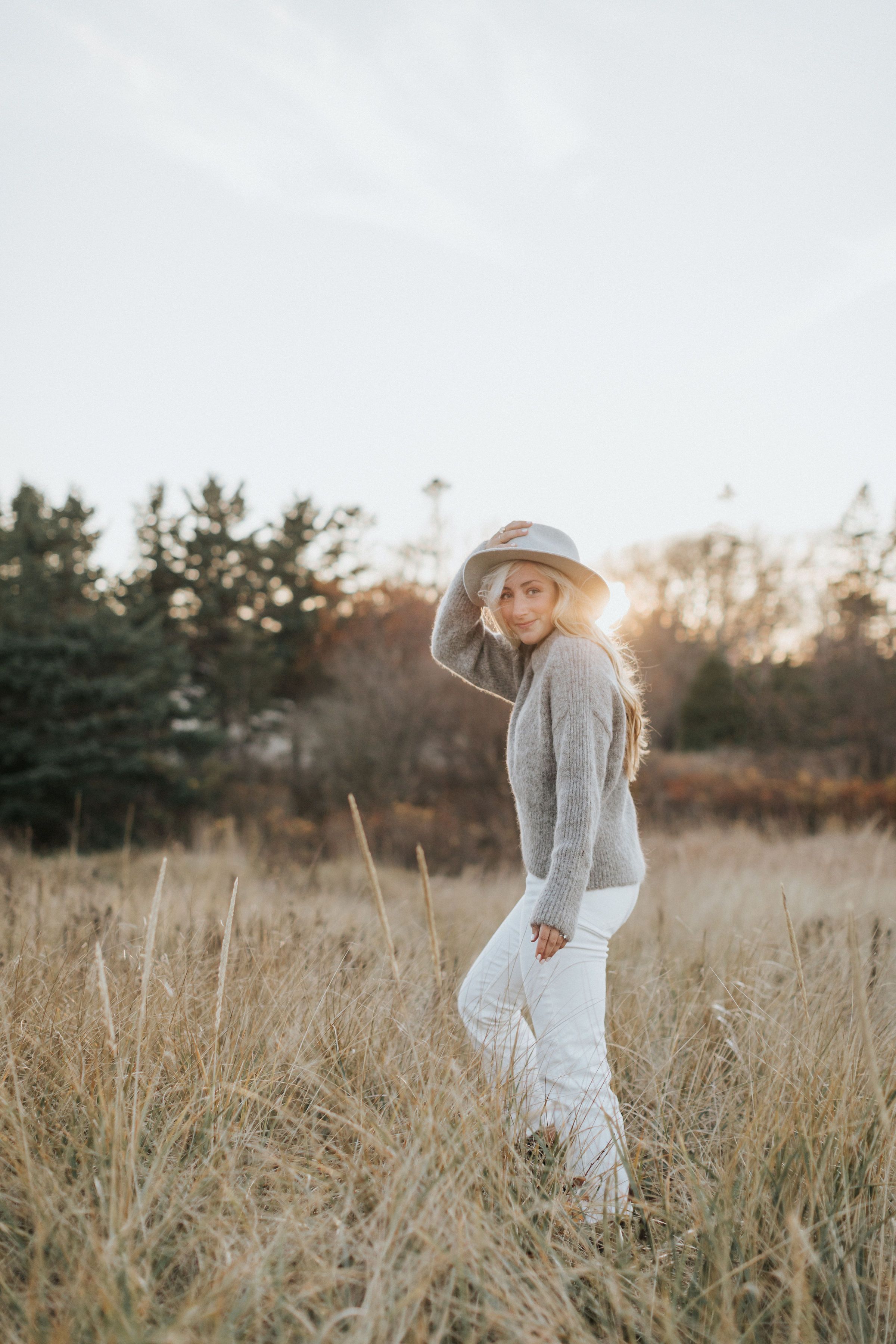 Person in grey sweater poses playfully in a golden sunlit field of tall grass.
