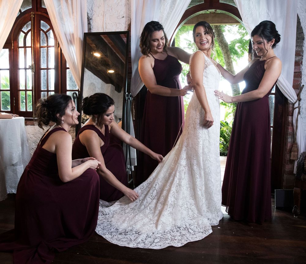 Bridesmaids in burgundy dresses helping adjust bride's lace wedding gown train.