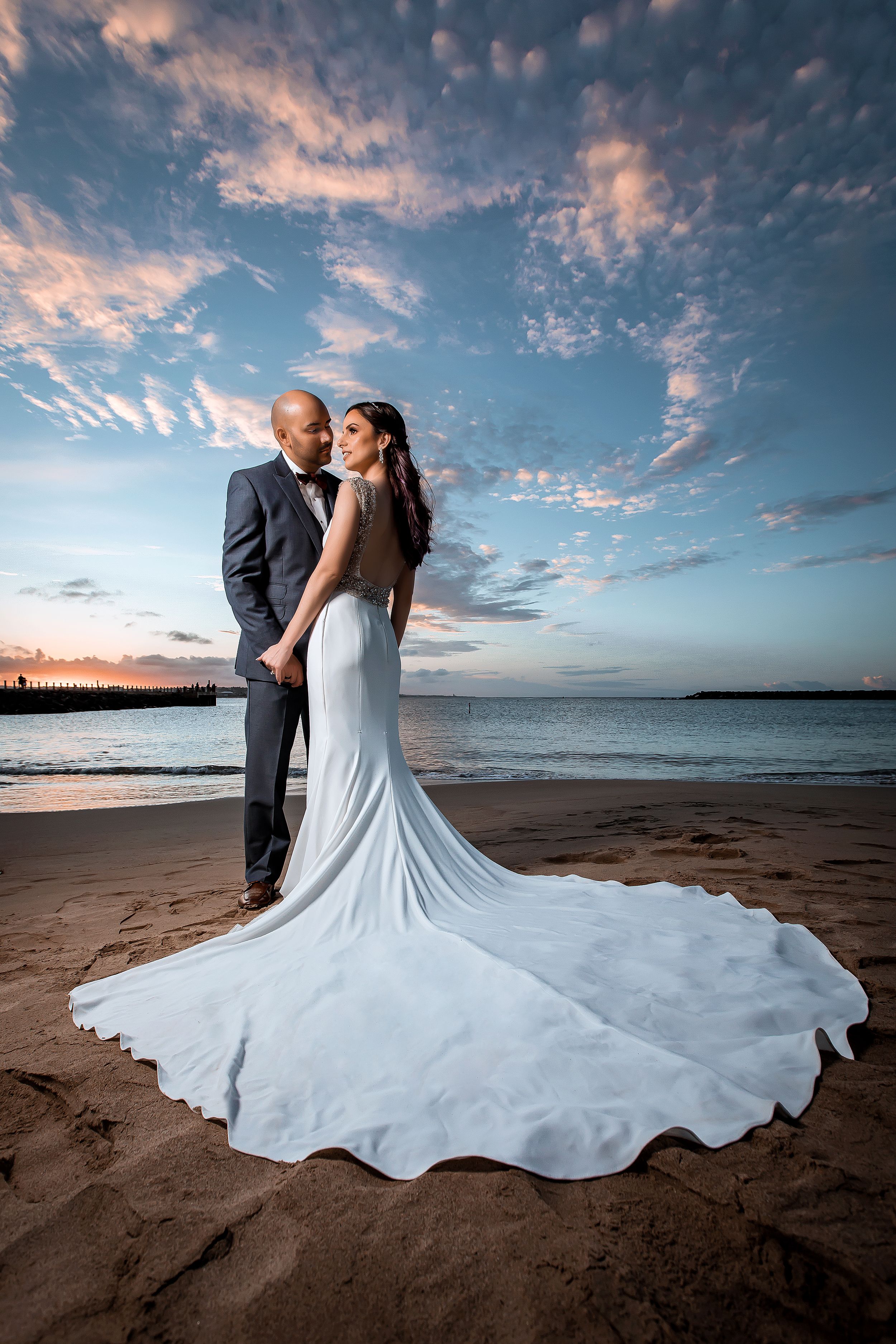 Dramatic beach wedding portrait with flowing dress against a colorful sunset sky.