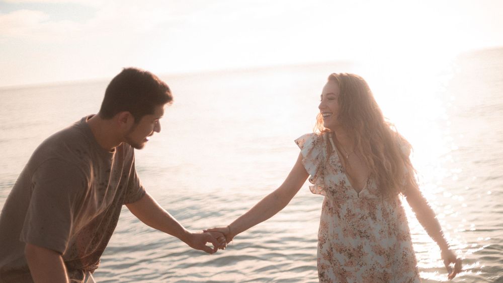 A couple holds hands while walking along the shoreline during sunset in a romantic seaside moment.