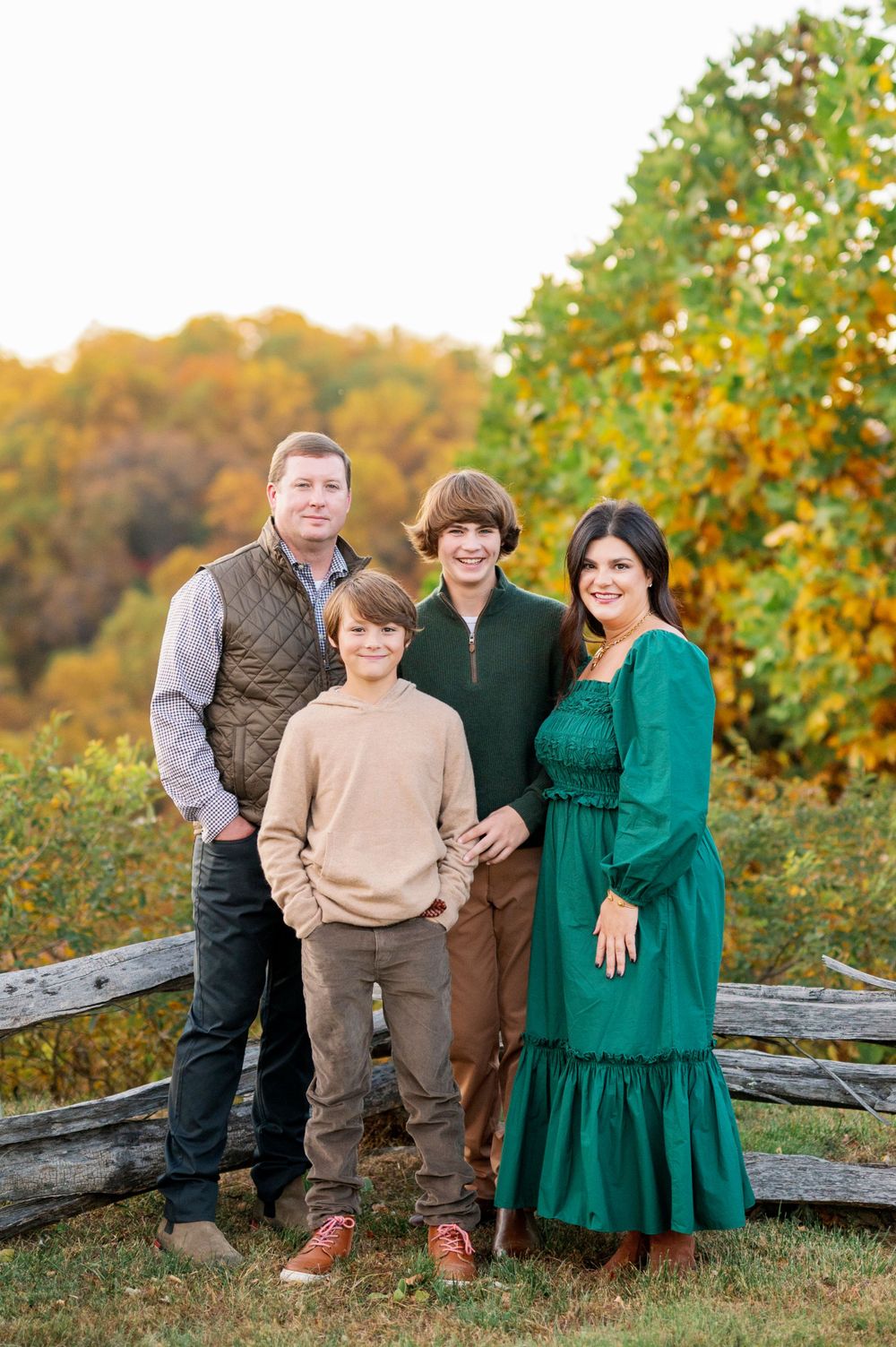 Family group poses together by rustic fence near autumn trees with orange leaves.