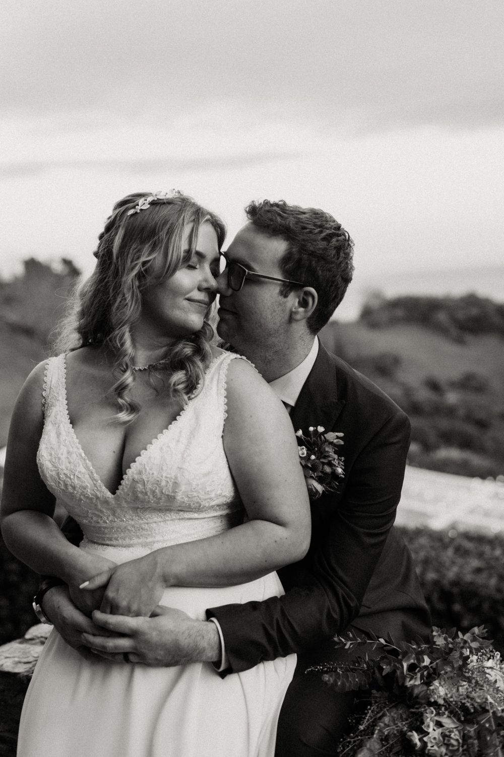 A romantic black and white wedding photo of a couple embracing outdoors with rolling hills in the background.