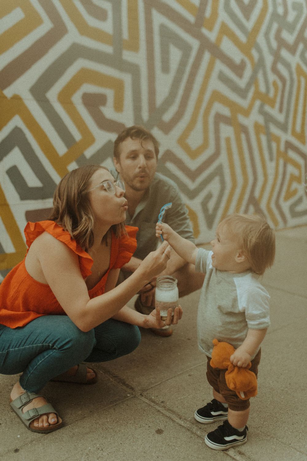 Family playing with bubbles together against a geometric patterned wall on a sunny day.