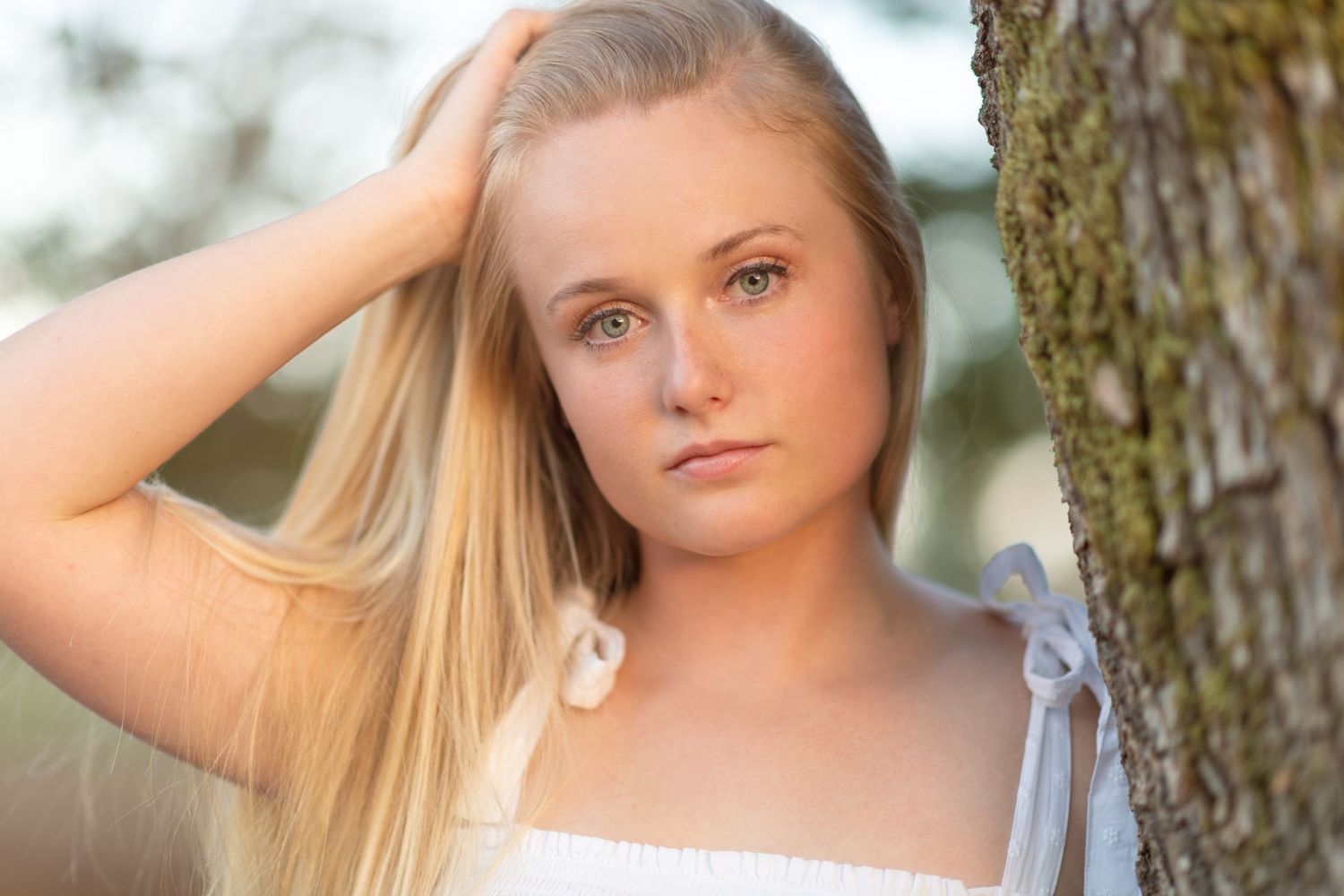 beautiful woman leaning shoulder against tree. hand in her blonde hair looking at camera.