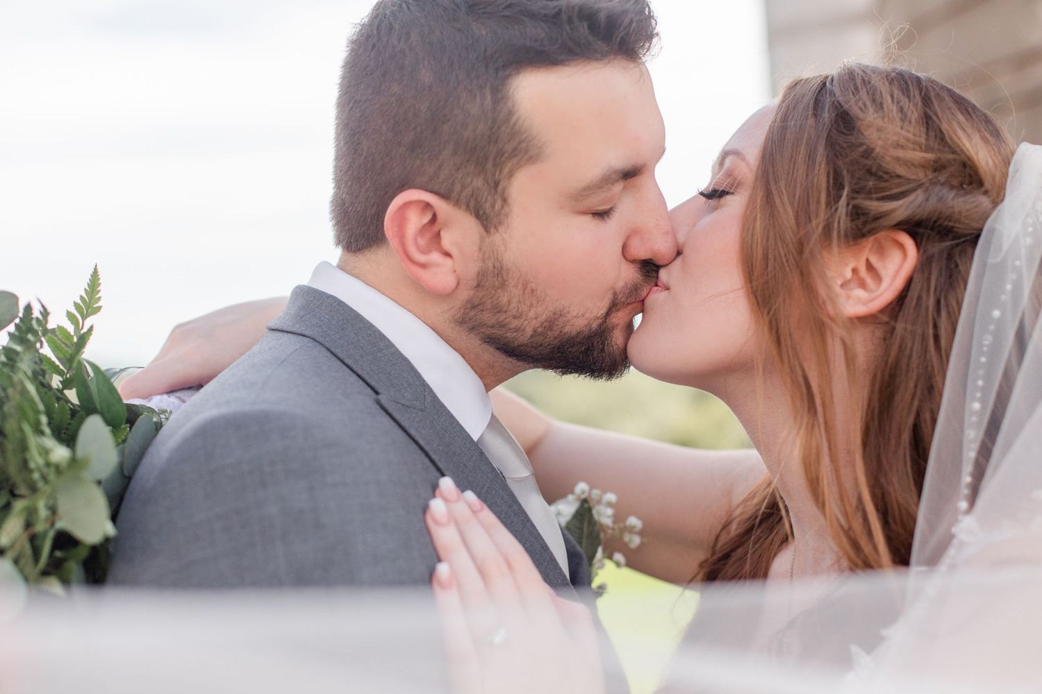 A tender wedding kiss moment captured through a delicate veil with greenery in the background.