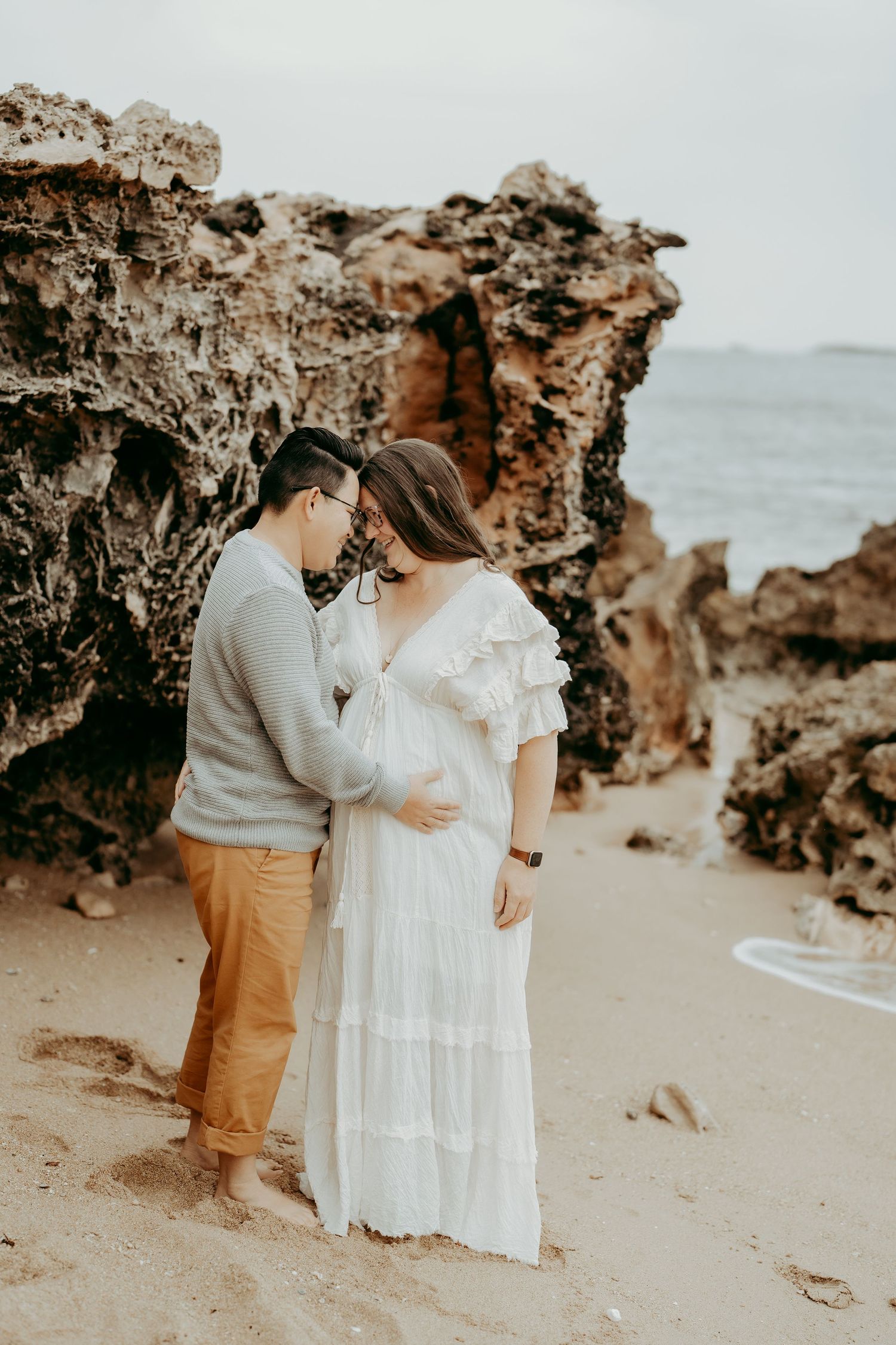 Couple embraces on beach with rocky cliffs in background during maternity photoshoot.