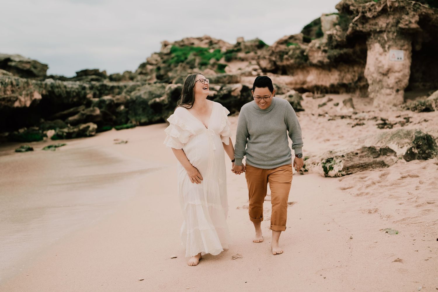 Couple walking and laughing together on beach with rocky cliffs behind them.