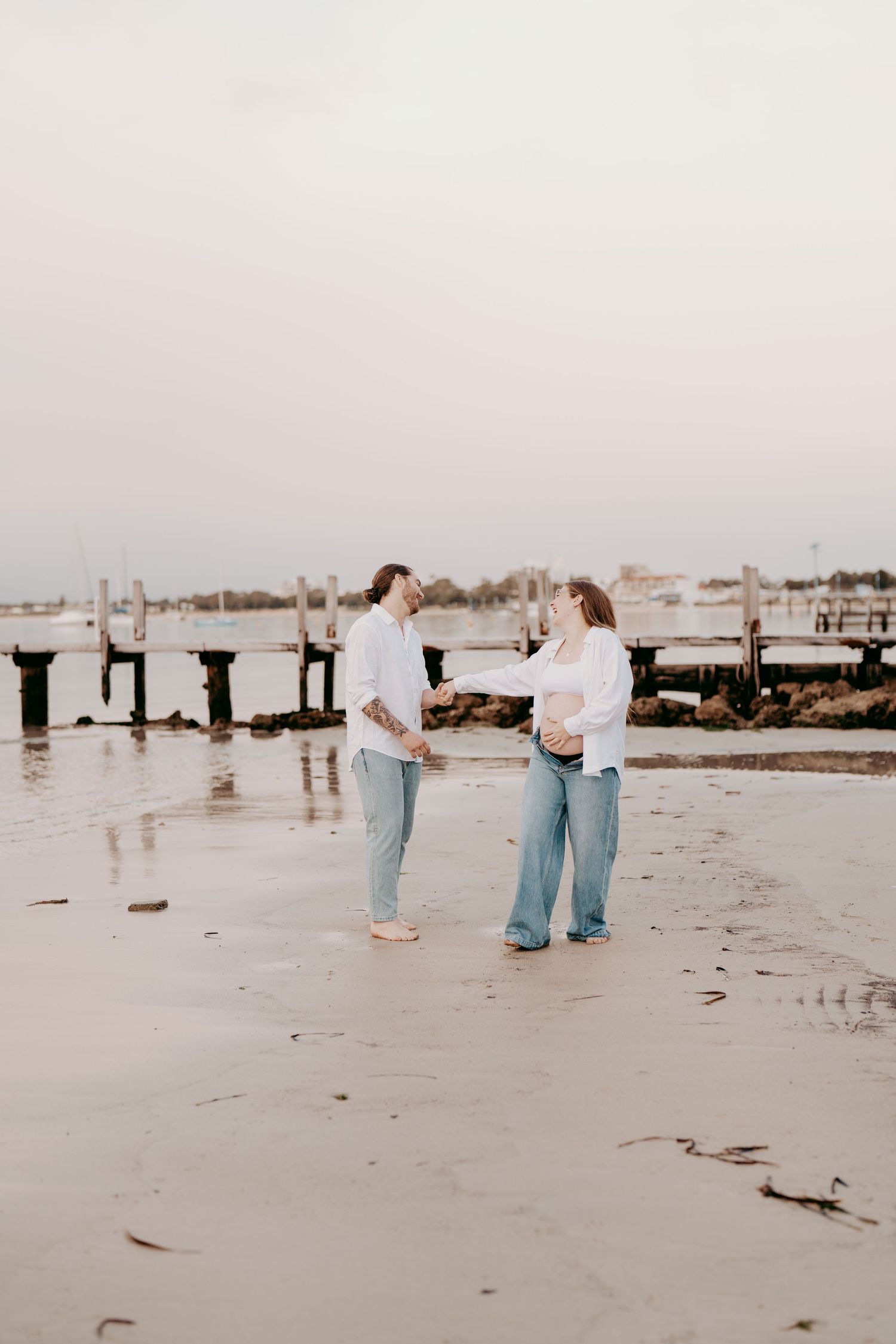 A couple in matching white shirts and jeans walking along a beach boardwalk at dusk.