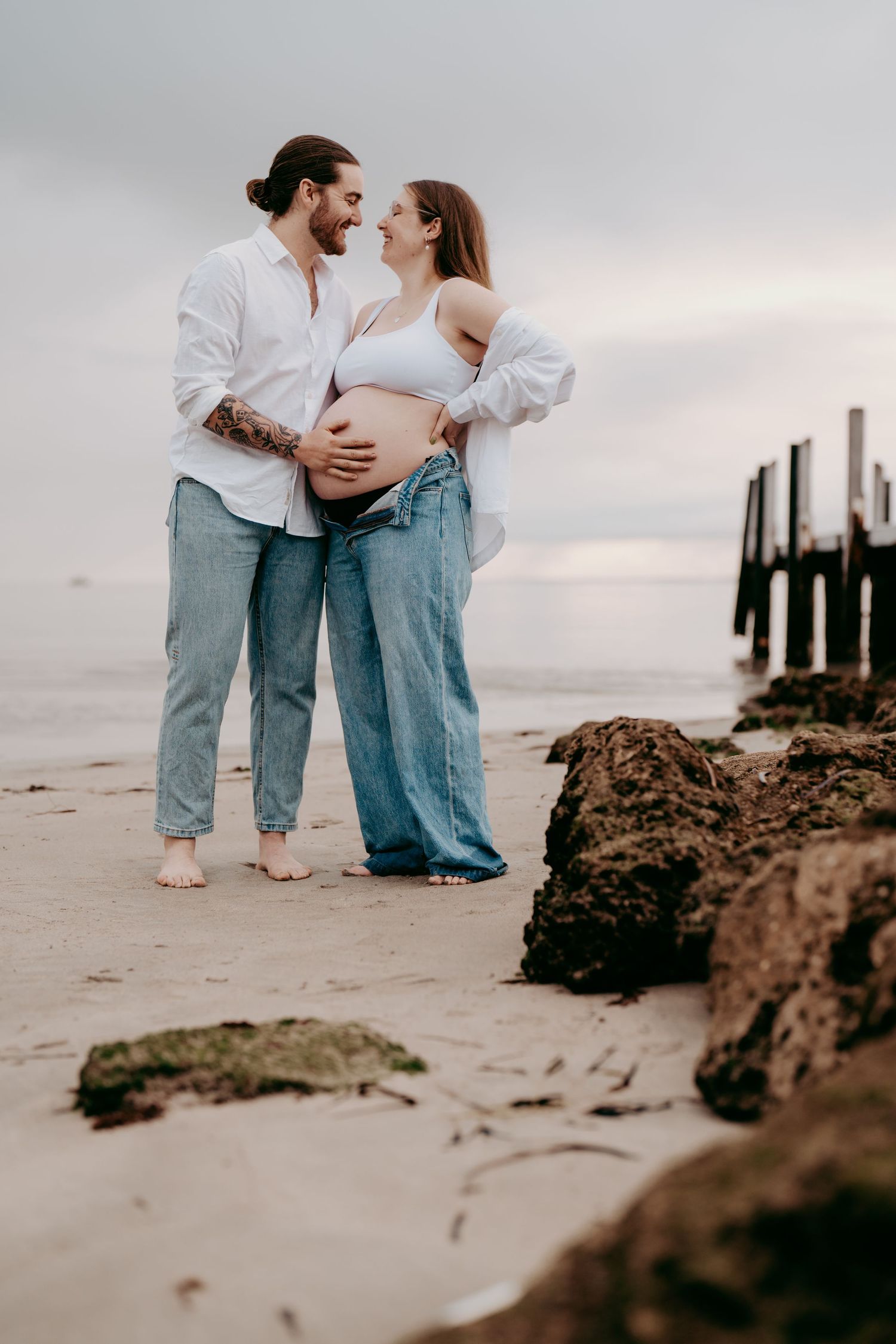 A romantic maternity photoshoot on a beach at sunset with an old pier in the background.
