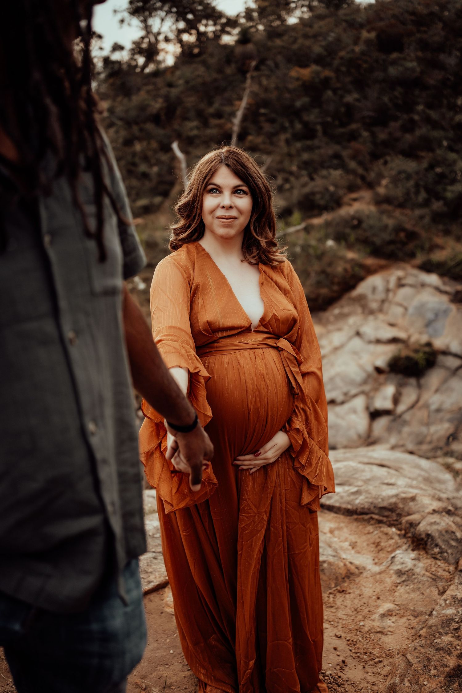 Maternity photo of woman in burnt orange dress standing on rocky terrain at sunset.