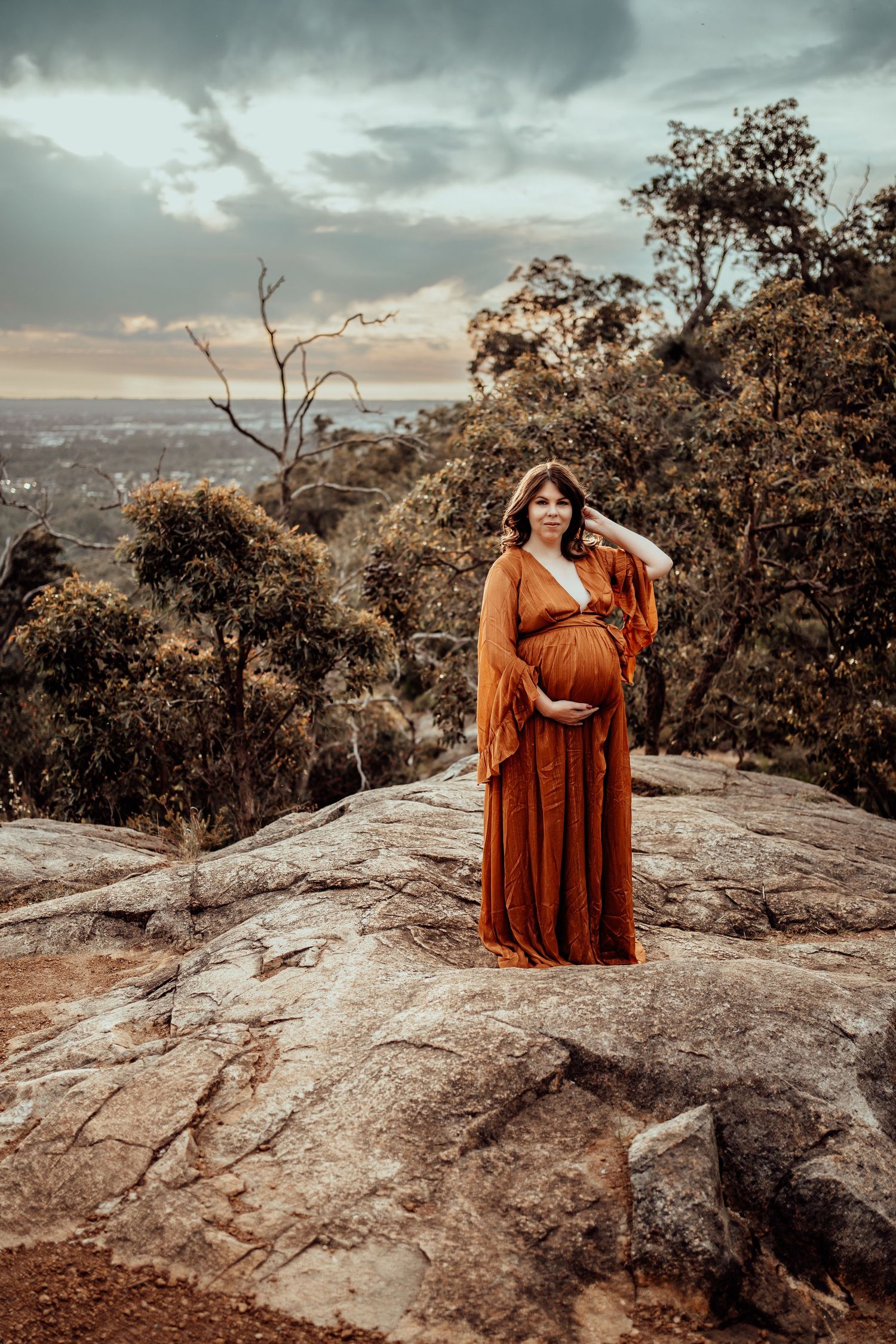 Pregnant woman in orange dress poses on rocky mountain overlook at sunset.