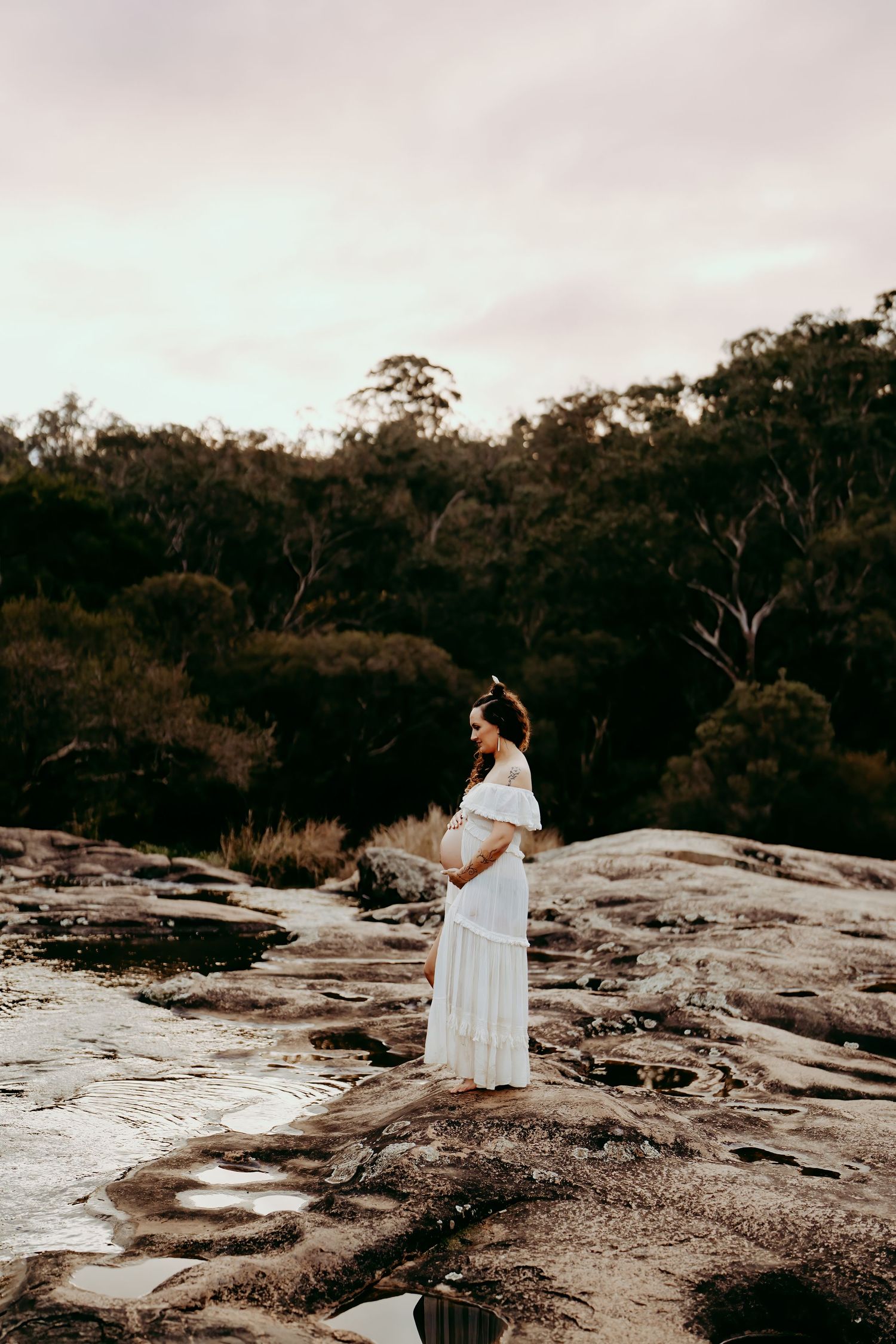 Expectant mother in white dress standing on rocky outcrop at dusk.