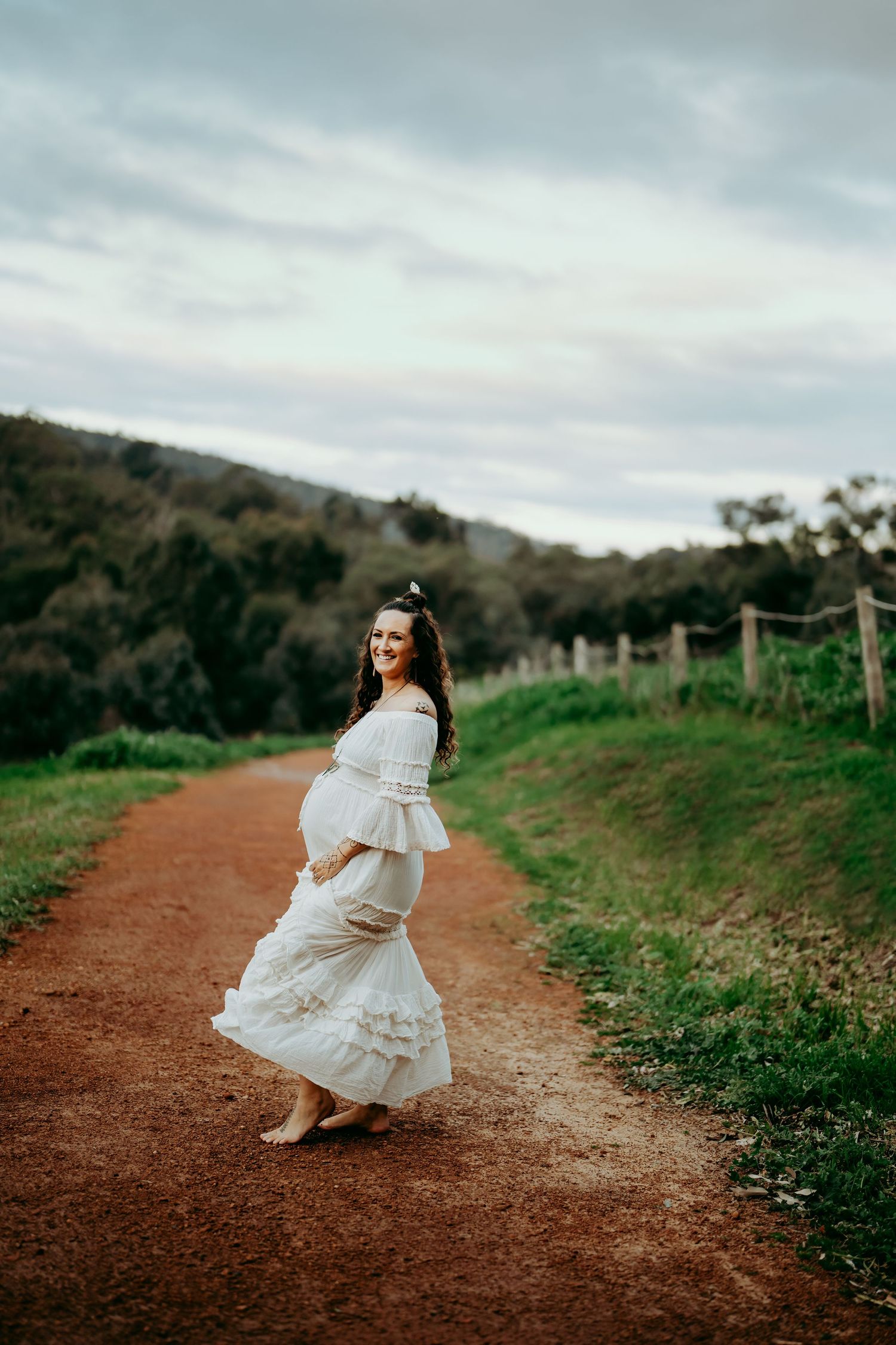 Pregnant woman in flowing white dress twirling on a rustic dirt path.
