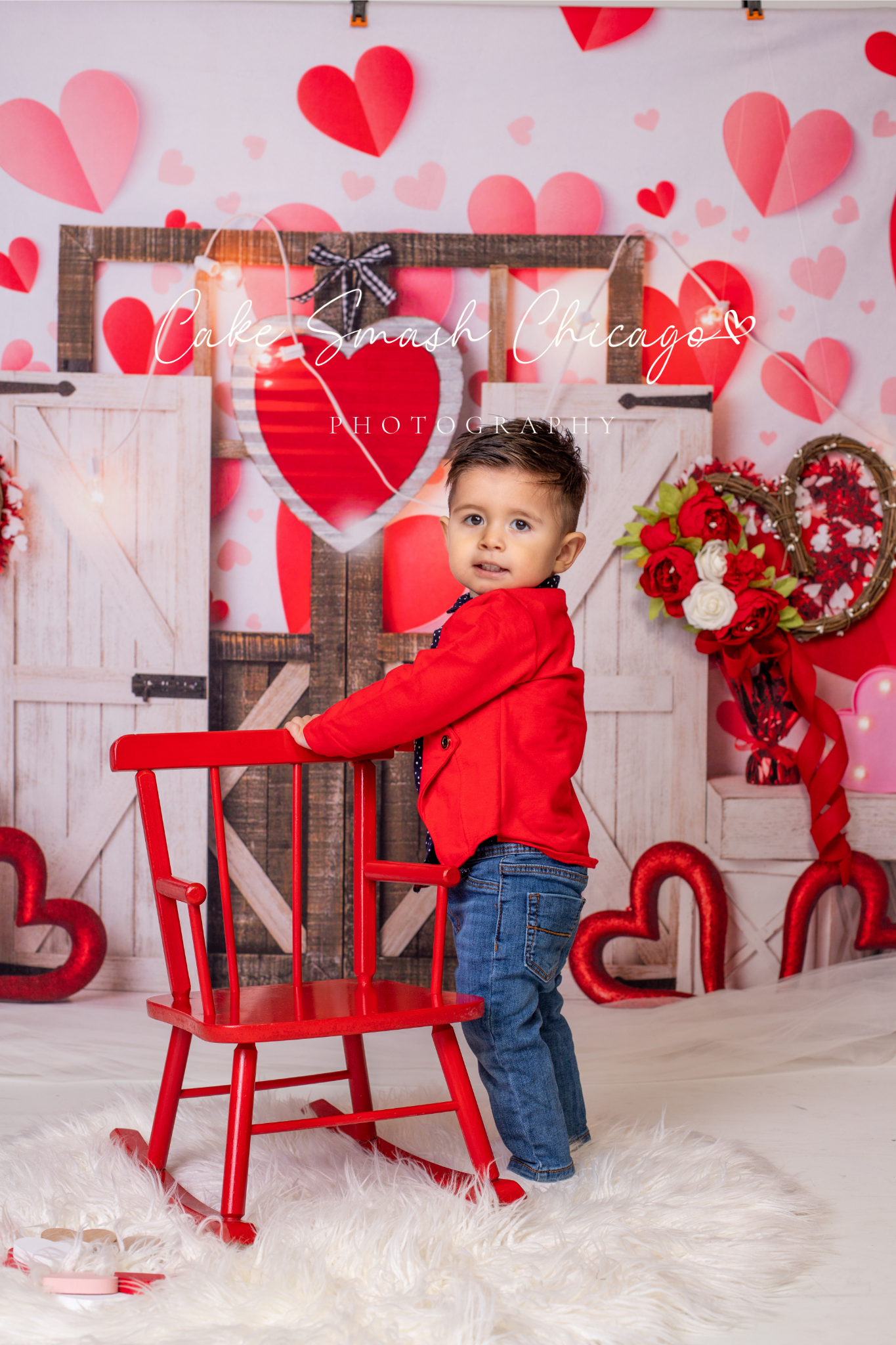 Valentine's Day themed photo setup with red rocking chair and heart decorations on white backdrop.
