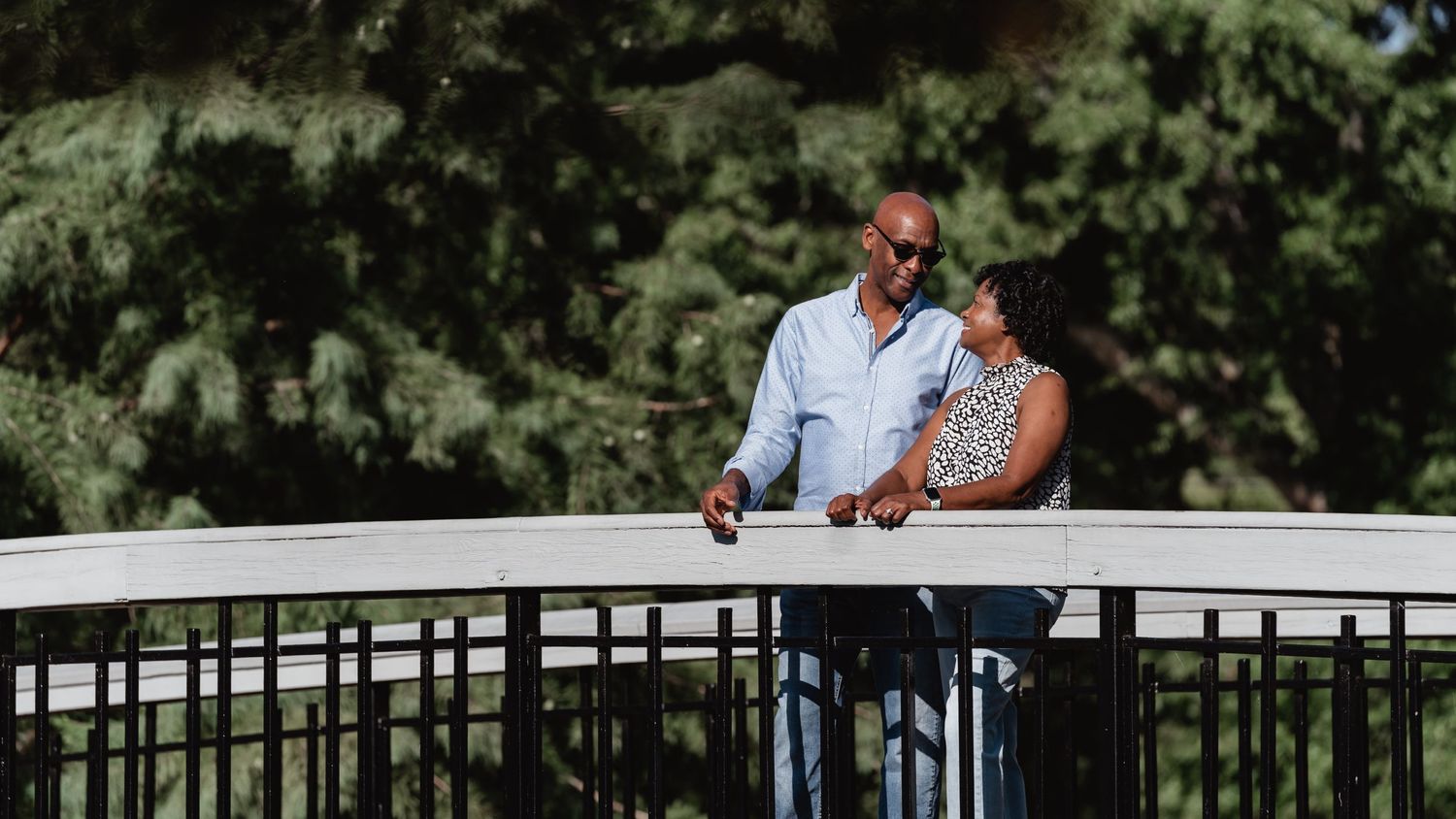 Couple standing together on bridge surrounded by green trees.