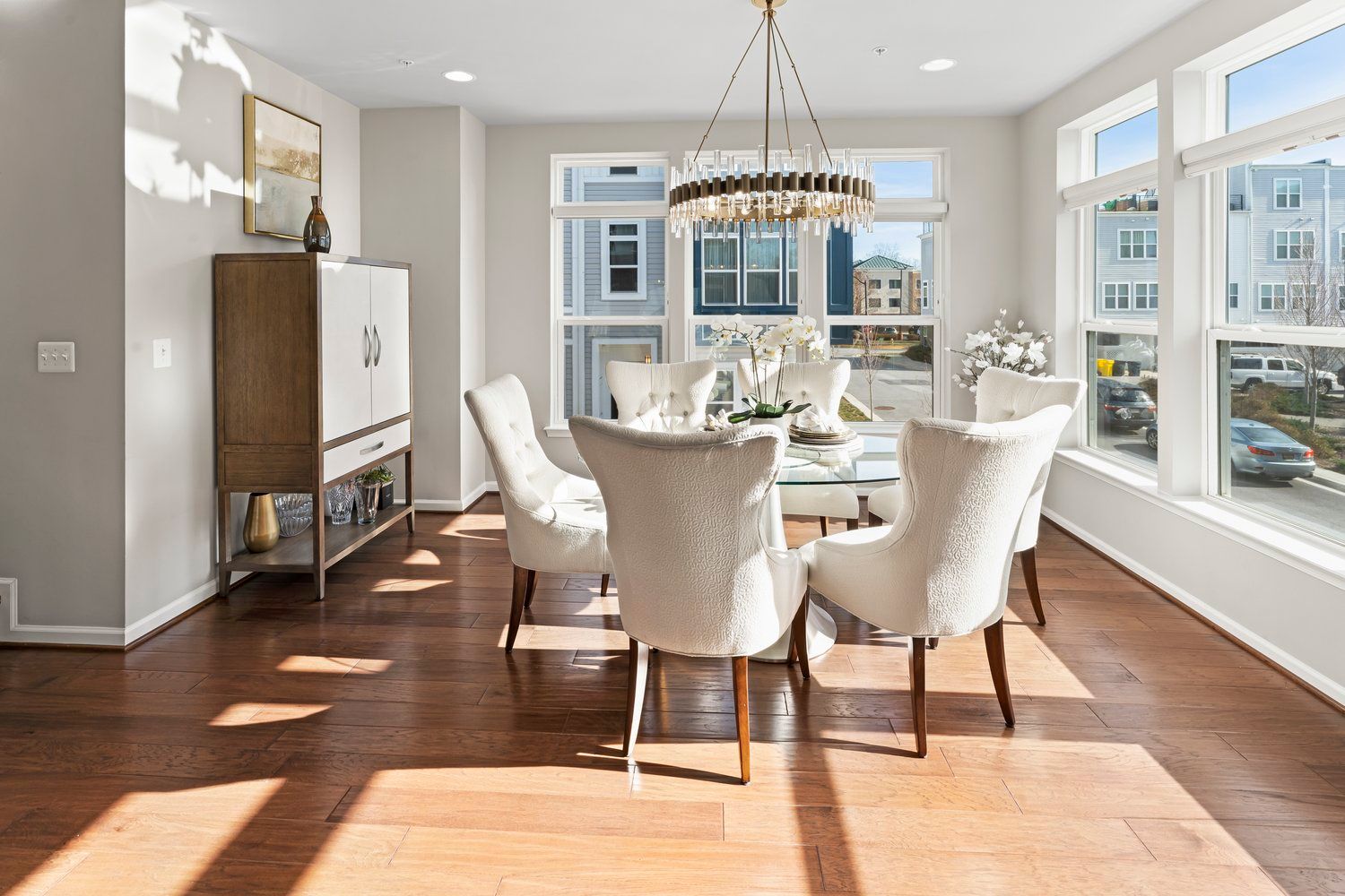Bright living room with large windows, hardwood floors, and elegant chandelier over dining area.