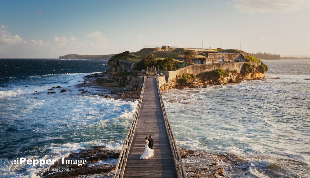 Wedding couple on wooden pier with dramatic ocean waves and historic fort backdrop.