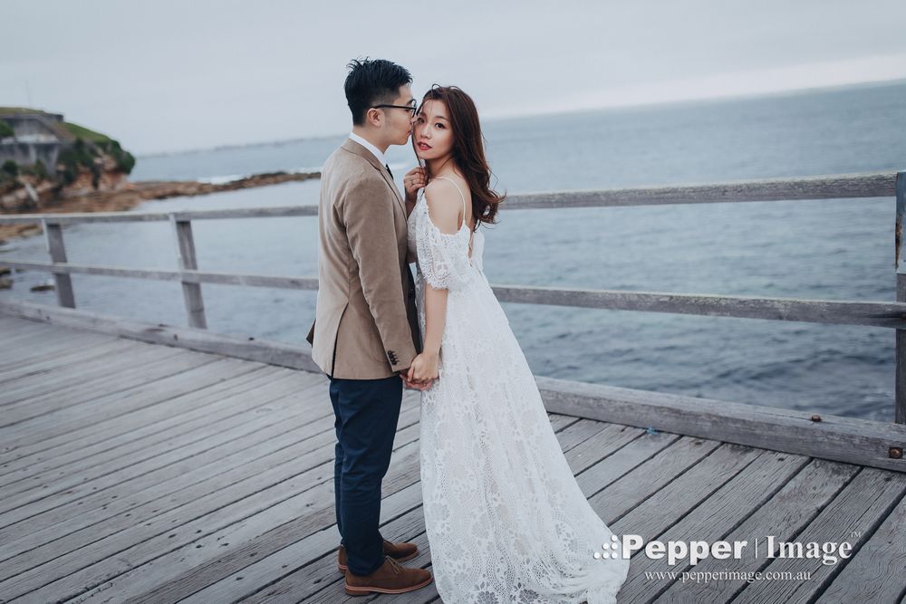 Couple holding hands on a wooden pier with calm ocean waters in the background.