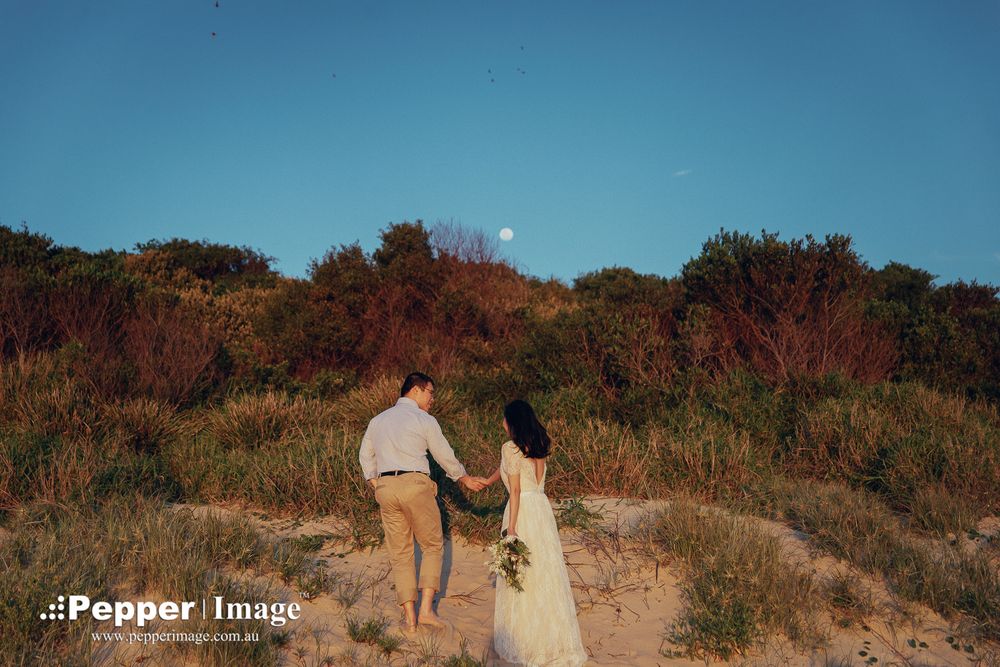 Couple walking hand-in-hand along a sandy dune path toward lush coastal shrubs at dusk.