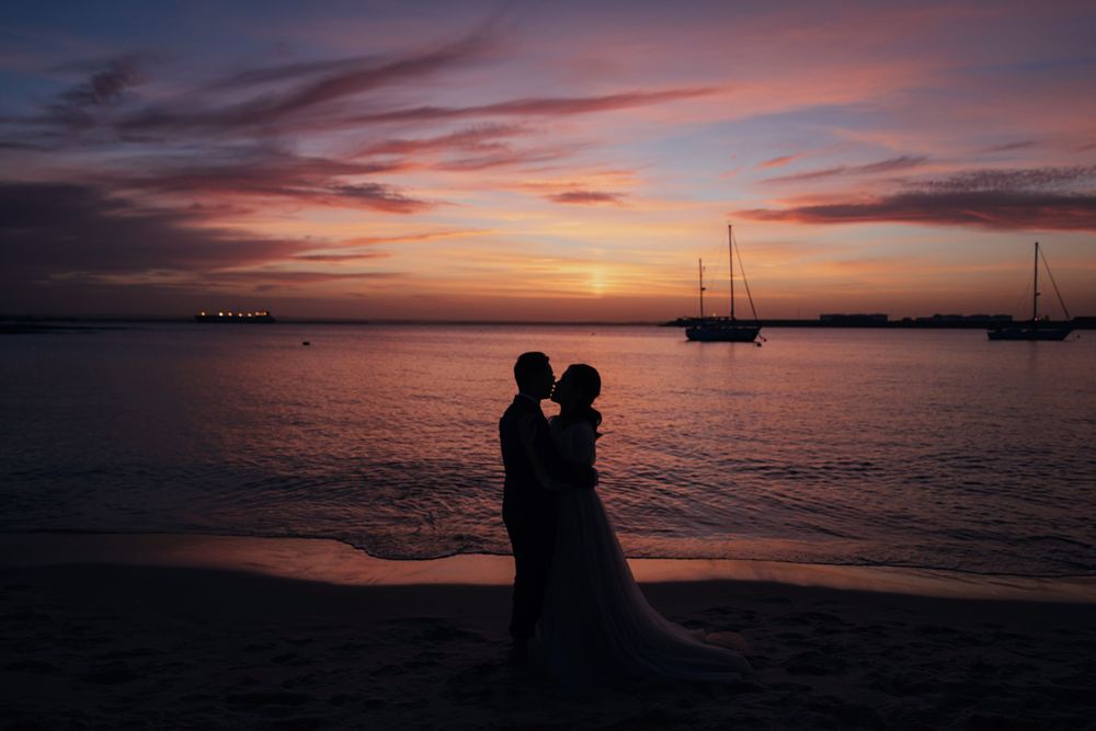 Romantic silhouette of wedding couple embracing on beach during vibrant purple sunset.