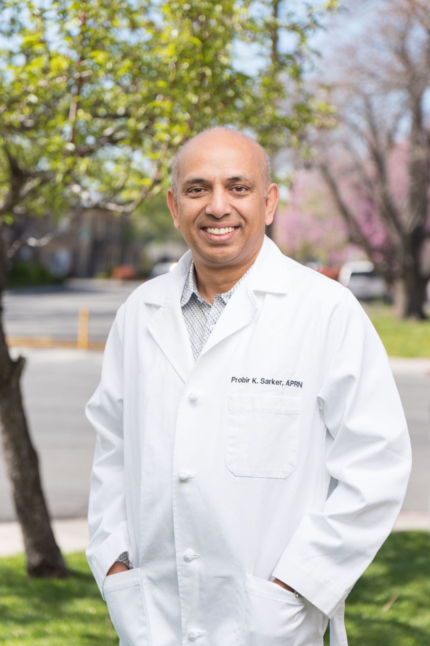 Healthcare professional in white lab coat stands outdoors with trees and greenery in the background.
