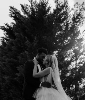 A romantic black and white wedding photo of a couple embracing under tall evergreen trees.