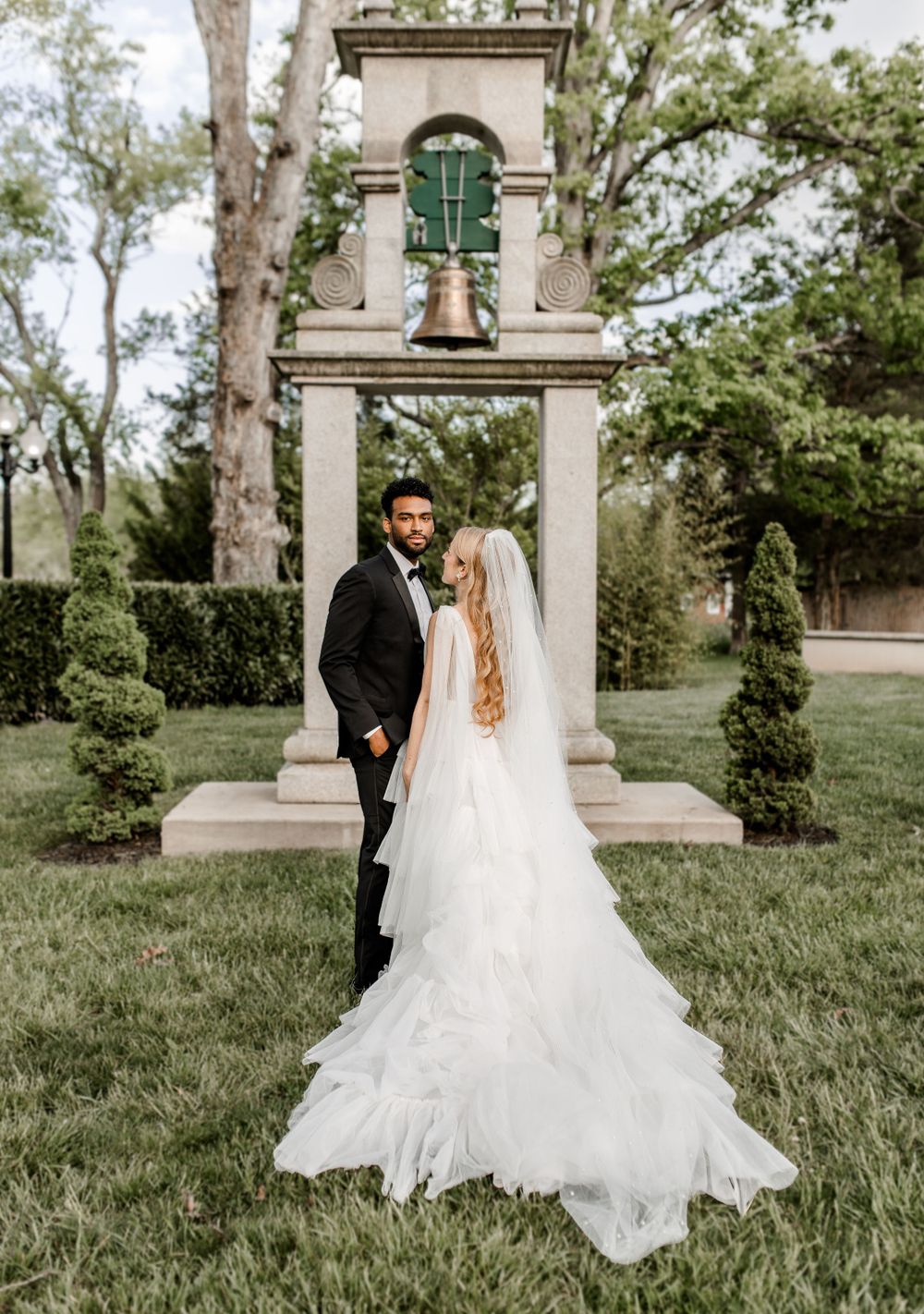 Wedding couple posing by ornate stone bell tower in garden setting.