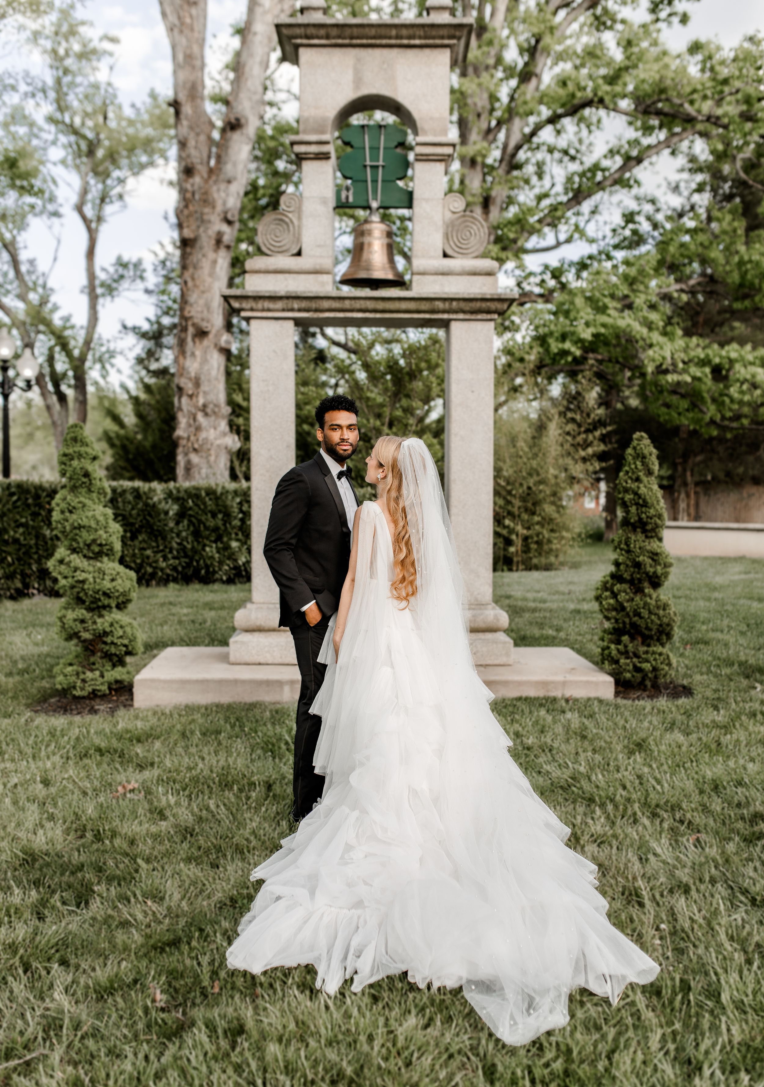 Wedding couple posing by ornate stone bell tower in garden setting.