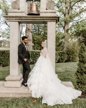 Wedding couple poses beneath an ornate stone bell tower surrounded by lush greenery.