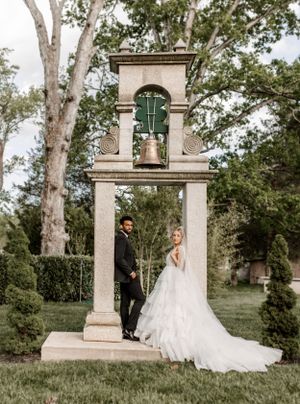 Newlywed couple poses elegantly by a stone bell tower structure in a garden setting.