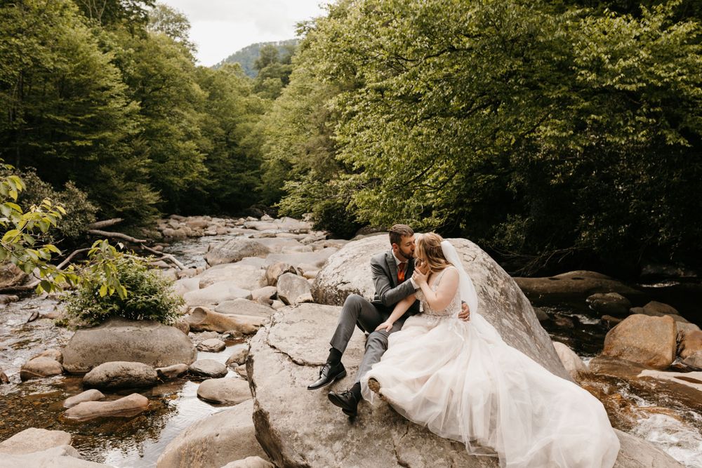 Bride and groom sit together on rocks by a mountain stream, sharing an intimate moment outdoors.