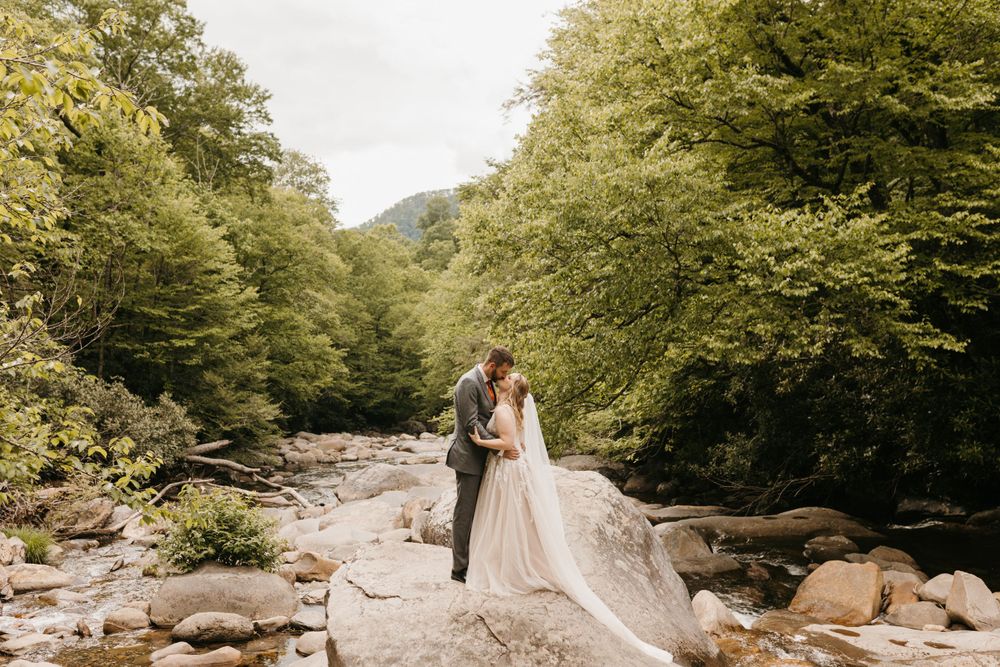 Bride and groom kiss on rocky riverbank surrounded by lush green trees and mountain scenery.