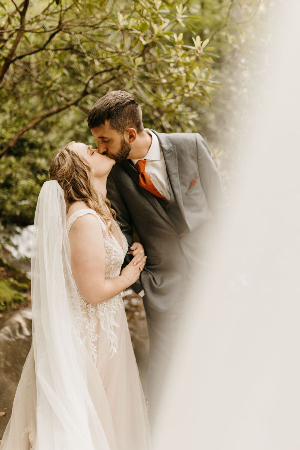 Bride and groom share a passionate kiss outdoors, groom wearing a gray suit with orange tie.
