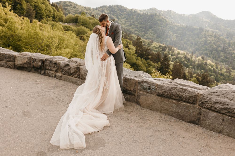 Bride and groom share a kiss as her long train flows on mountain overlook path.