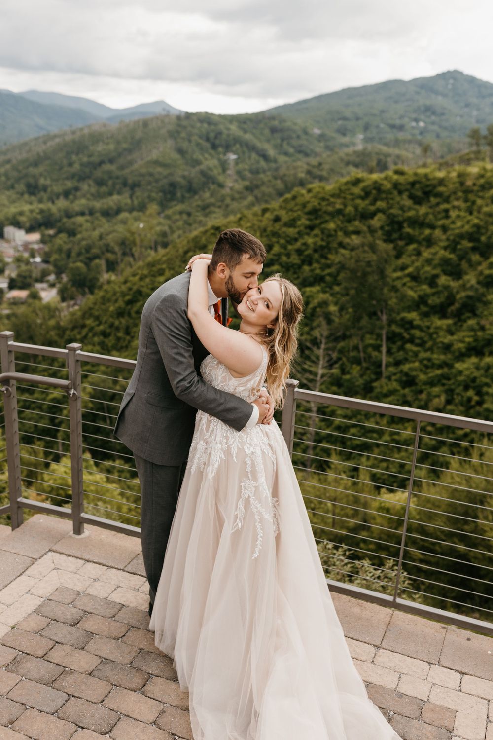Groom kisses bride on the cheek as she smiles on a mountain terrace with green hills behind them.