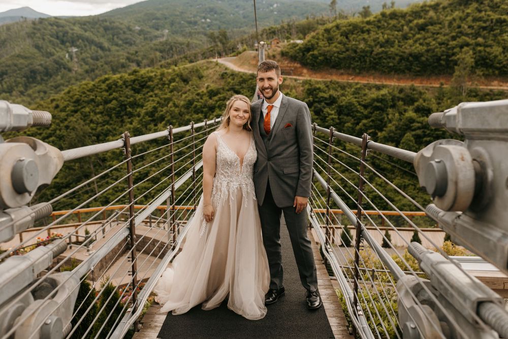 Couple in wedding attire posing on a suspension bridge with mountain views.