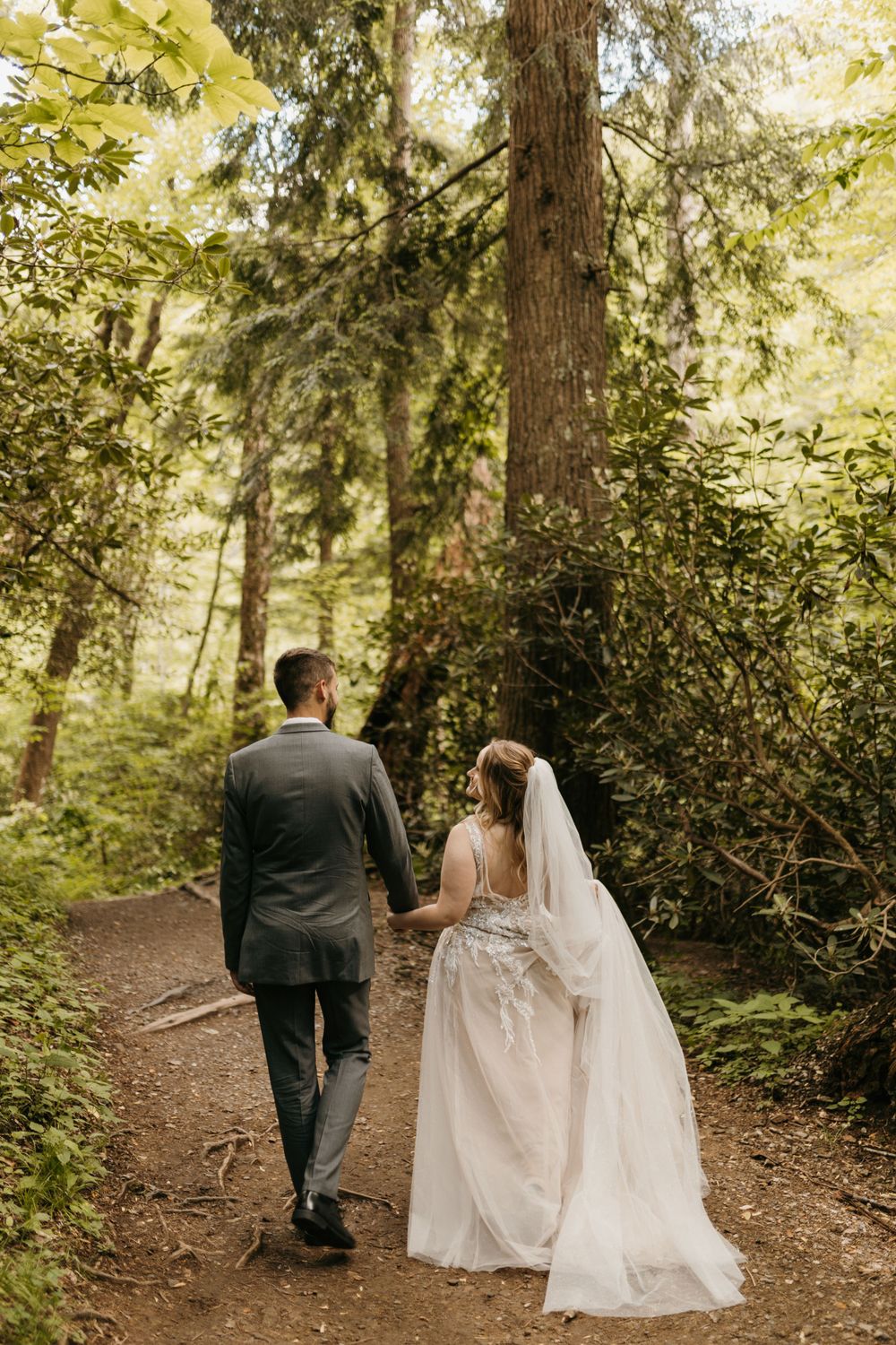 Bride and groom walk hand in hand down a forest path, bride wearing a flowing white gown with veil.
