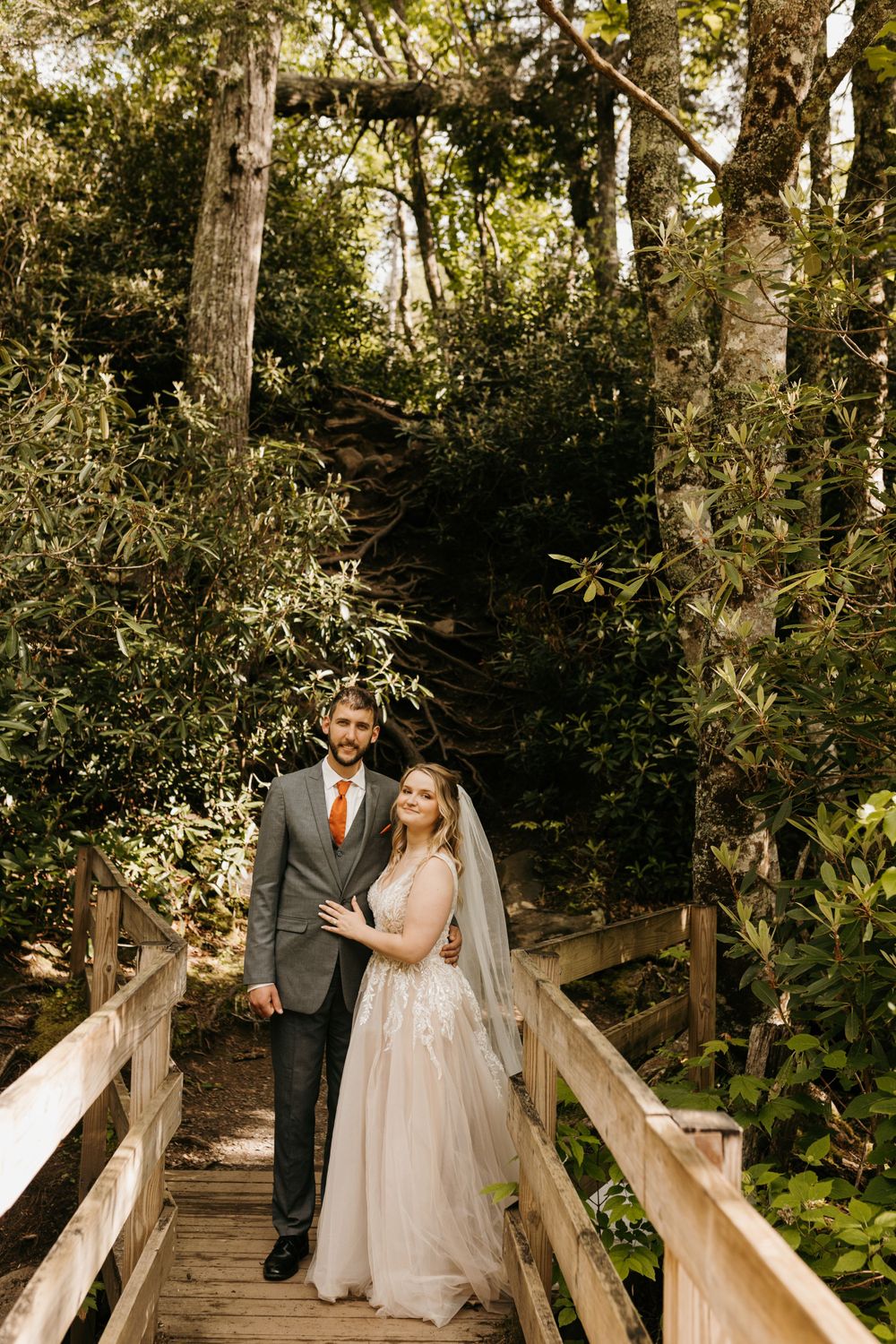 Bride and groom stand together on a wooden bridge surrounded by dense green forest foliage.