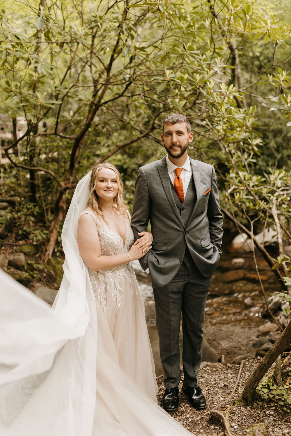 Smiling bride and groom pose together in a lush forest setting, groom wearing an orange tie.
