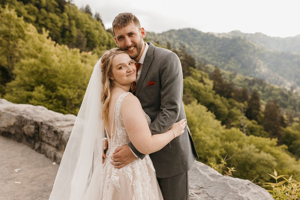 Smiling bride and groom embrace at stone overlook surrounded by lush mountain scenery.