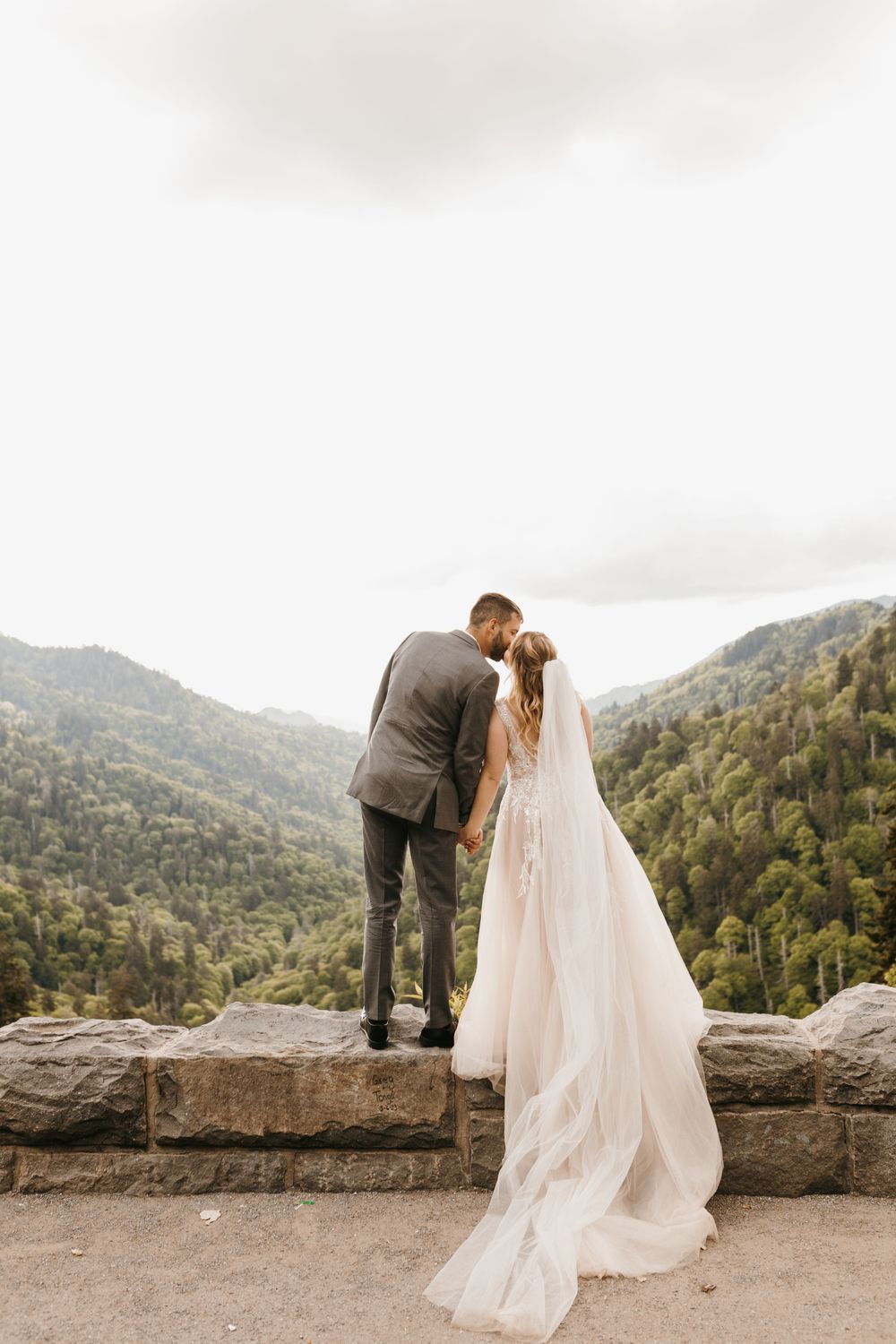 Groom kissing bride on forehead at scenic mountain overlook with flowing veil.
