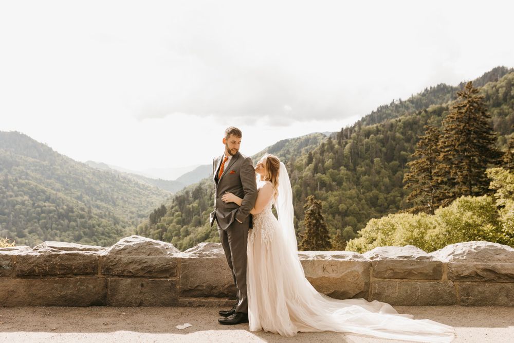 Bride and groom share a tender moment at stone wall overlook with mountain views.