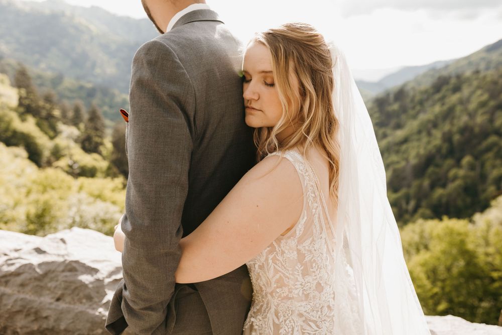 Bride in lace veil embraces groom from behind with lush mountain forest backdrop.