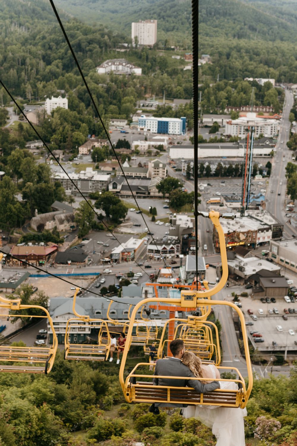 Bride and groom ride a yellow ski lift chairlift high above a scenic mountain resort town.