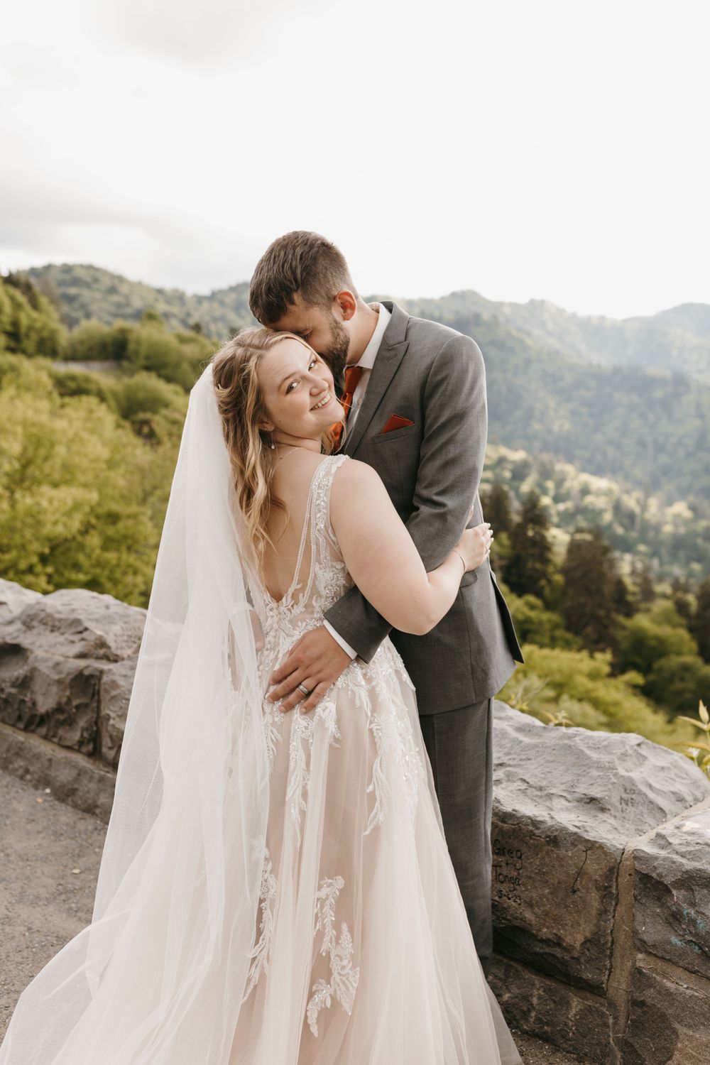 Groom nuzzles smiling bride in lace wedding gown against scenic mountain backdrop.