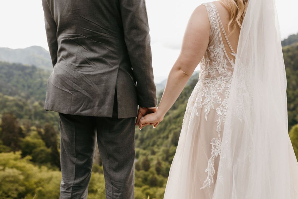 Close-up of bride and groom holding hands with mountain greenery behind them.