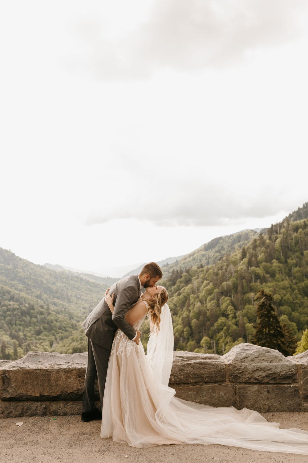 Groom kisses bride as her flowing gown trails on mountain overlook at elopement.