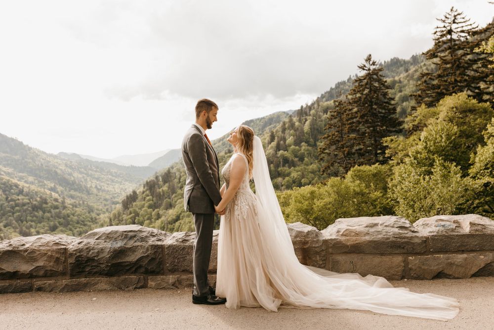 Bride and groom hold hands facing each other on a stone overlook with mountain views behind them.