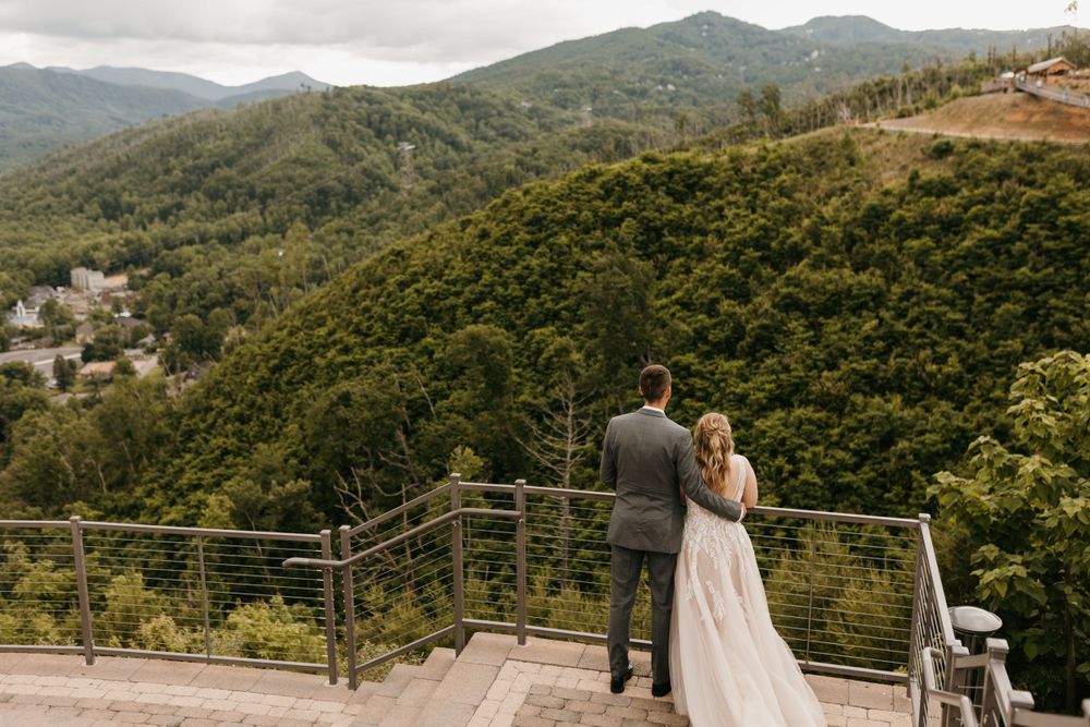 Bride and groom stand together on a scenic mountain overlook deck, gazing at lush green hills below.